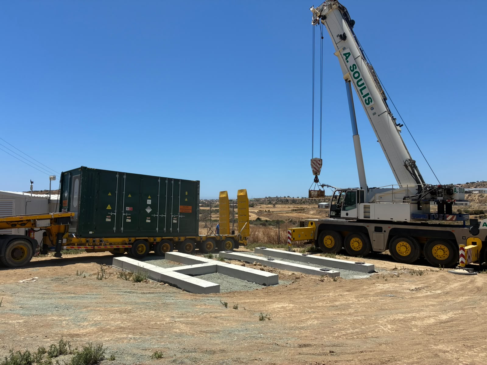 Battery container on a delivery trailer beside a crane and prepared concrete foundations